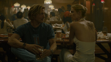 A man and woman sit at a bar table talking, each holding beer mugs. The setting is a dimly lit, busy restaurant or bar with other patrons in the background and a casual atmosphere.