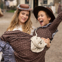 Two girls in vintage dresses and hats, one carrying the other while laughing, outdoors in what appears to be a historical or rural village setting with blurred buildings and people in the background.