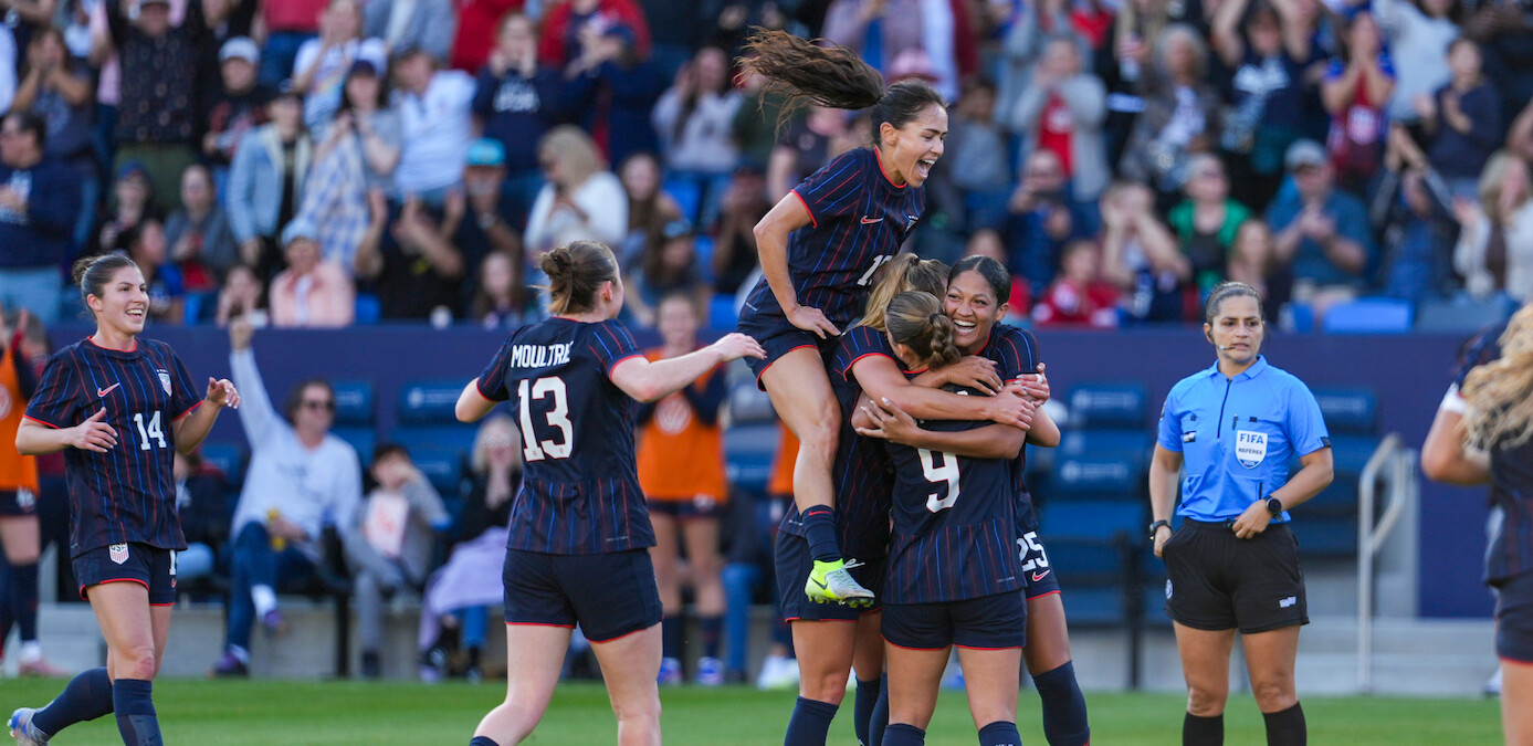 Women's soccer team celebrates a goal on the field; players embrace and jump with joy while a referee and cheering crowd are visible in the stadium background.