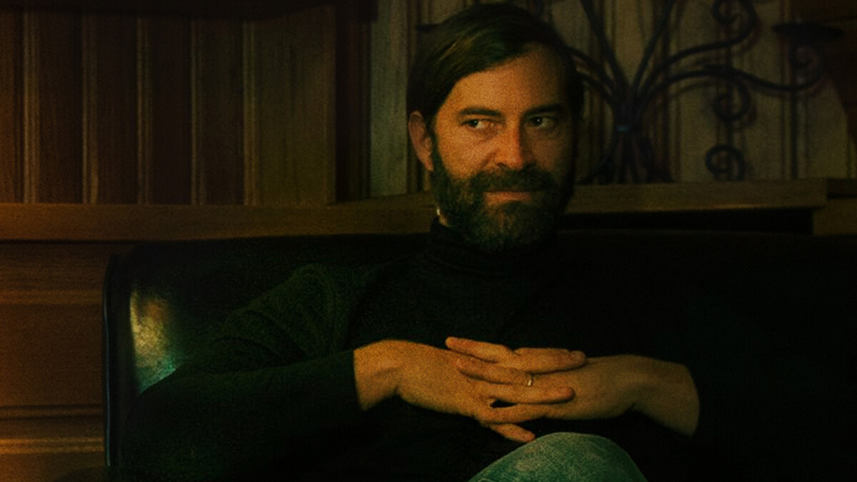 Bearded man with dark hair sits on a black leather chair in a warmly lit wooden room, wearing a black turtleneck and jeans, hands folded, looking to the side with a thoughtful or mischievous expression.