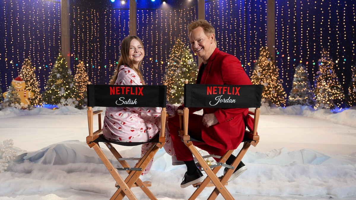 Two people smiling while sitting on director’s chairs with Netflix logos in a festive winter set decorated with snow, Christmas trees, twinkling lights, and holiday decorations, creating a cheerful holiday atmosphere.