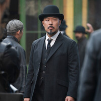 A serious man in a black pinstripe suit and bowler hat stands in an urban street scene, surrounded by others in period clothing, with green and yellow building windows in the background.