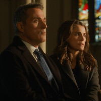 A man in a suit and a woman in a dark coat sit side by side in a dimly lit church, with stained glass windows in the background, both looking thoughtful and serious.
