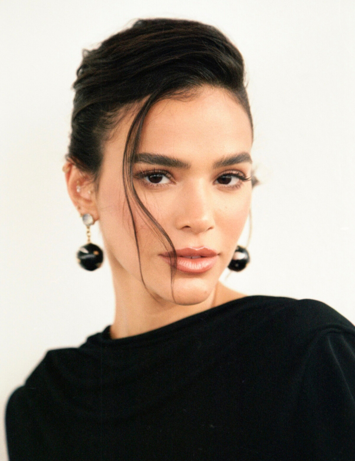 Bruna Marquezine é uma mulher branca, magra, de cabelos escuros e presos, com brincos grandes e veste um vestido preto