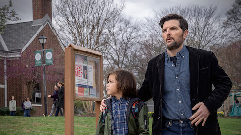 A man and child stand outside near a community notice board with a "Have You Seen Me?" poster. They look alert or concerned. The setting is a grassy area by a brick building with people and leafless trees in the background.