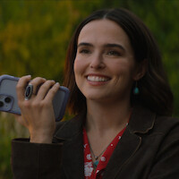 Smiling woman with dark hair holding a phone outdoors, wearing a brown jacket, red shirt, turquoise jewelry, and standing in front of a blurred green background.