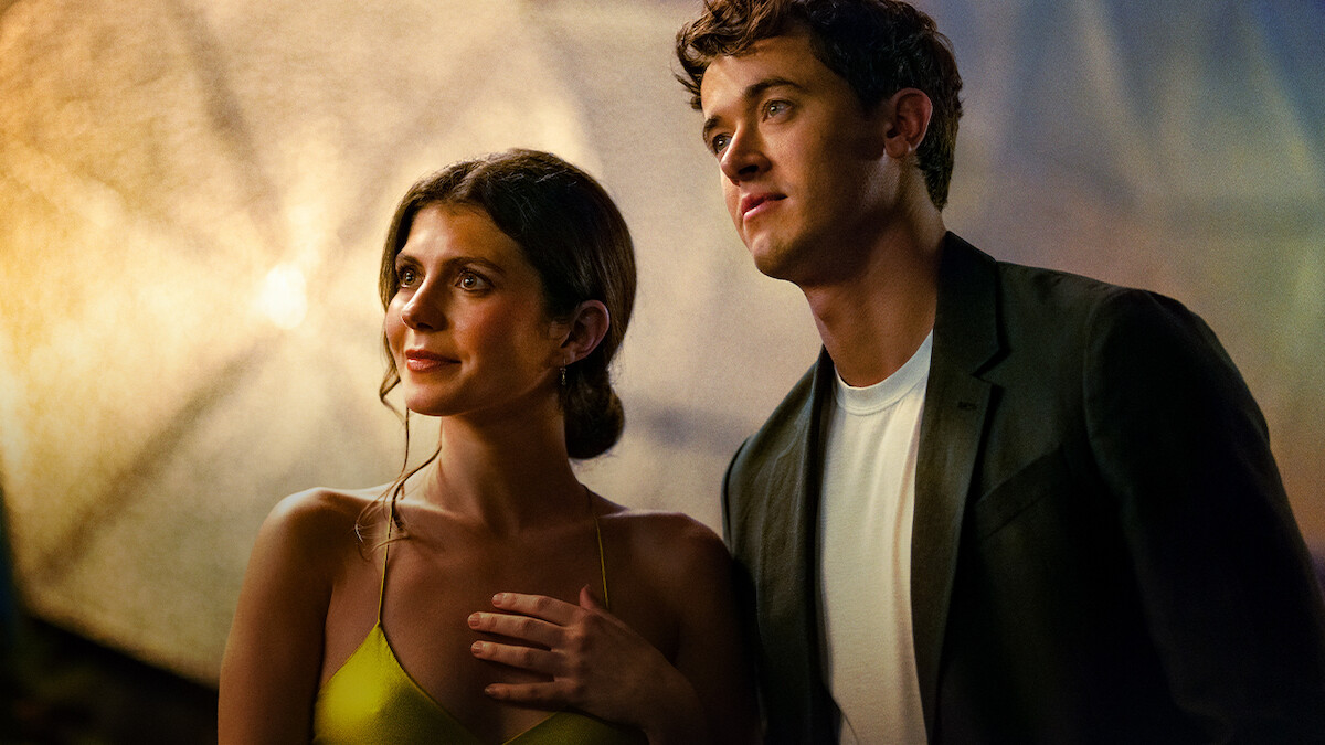 A woman in a yellow dress and a man in a dark blazer stand close together under warm, soft lighting, looking off into the distance.
