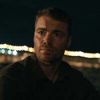 A man with short hair and a trimmed beard sits outdoors at night, illuminated by soft lights in the background, creating a pensive atmosphere with blurred string lights and dark sky.