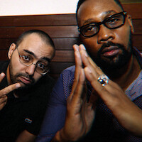 Two men sitting indoors against a wooden wall, one wearing glasses and making a peace sign, the other in a blue shirt and glasses with hands clasped together, both facing the camera in a casual setting.