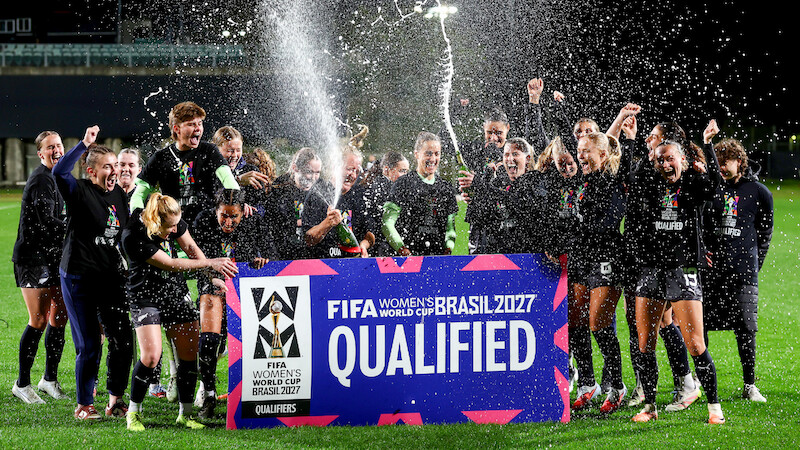 Women's soccer team celebrates at night on a field, spraying champagne and cheering behind a large sign reading "FIFA Women's World Cup Brasil 2027 Qualified." Bright stadium lights and festive atmosphere.