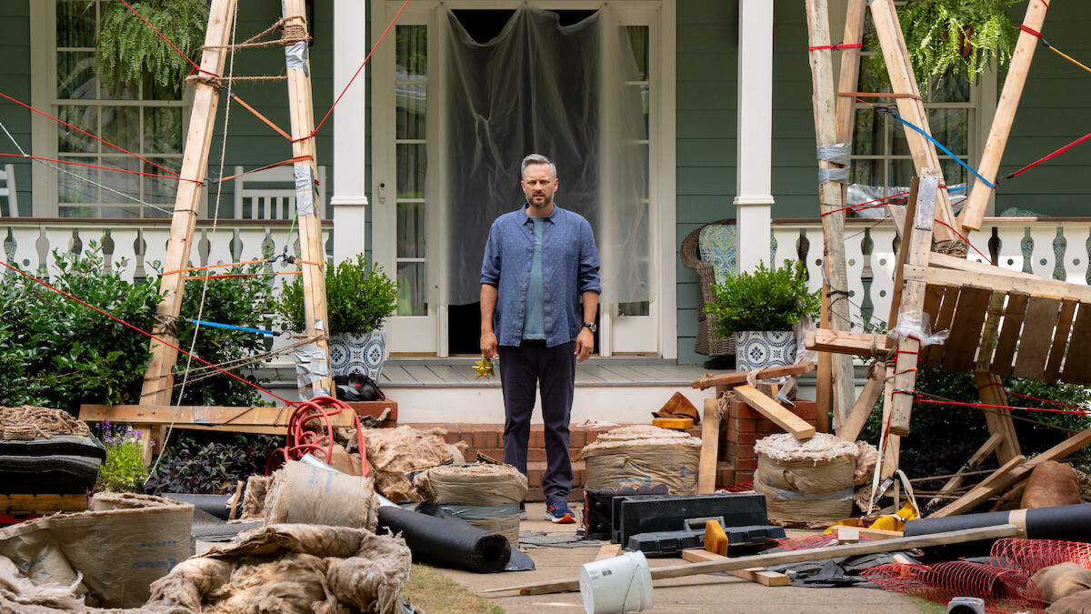 A man stands on a porch surrounded by construction materials, debris, and wooden scaffolding in front of a house. The setting suggests renovation work in progress, with scattered tools and building supplies.