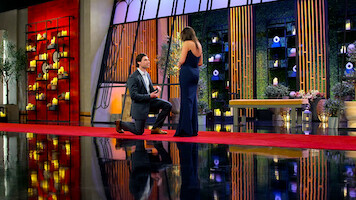 A man kneeling to propose to a woman in an elegant indoor setting with red carpet, decorative lighting, plants, and flower arrangements, creating a romantic atmosphere.