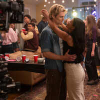 A young couple dances closely at a lively house party with friends in the background, red cups on a table, string lights overhead, and a film crew camera partially visible, capturing the casual atmosphere.