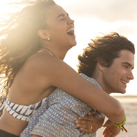 A joyful young woman laughs while riding piggyback on a smiling young man at the beach during sunset, with sunlight creating a warm, cheerful atmosphere.