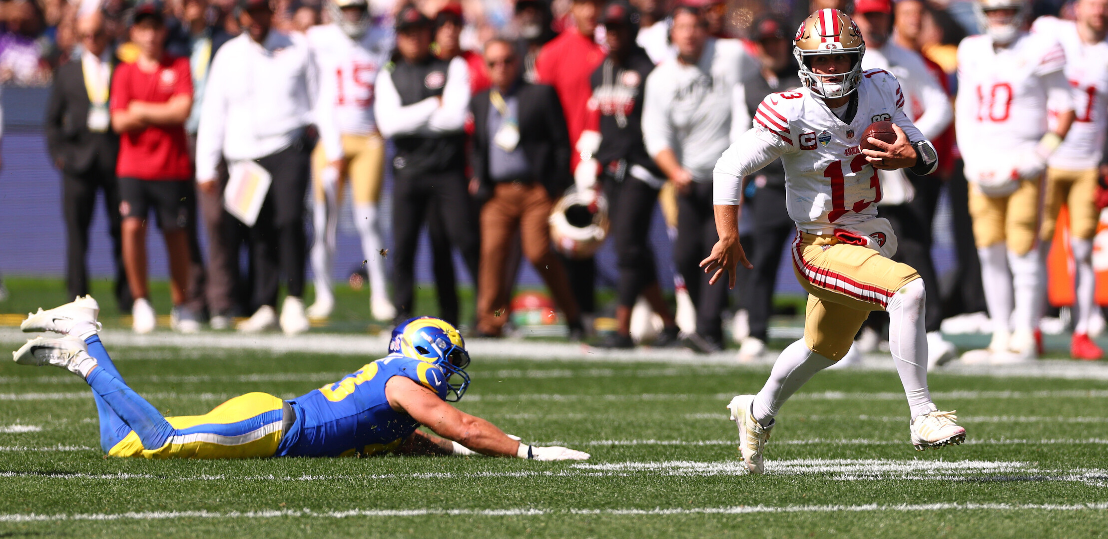 Football quarterback in a white and gold uniform runs with the ball on a sunny field while a defender in a blue and yellow uniform dives to tackle him; crowd and teammates watch in the background.