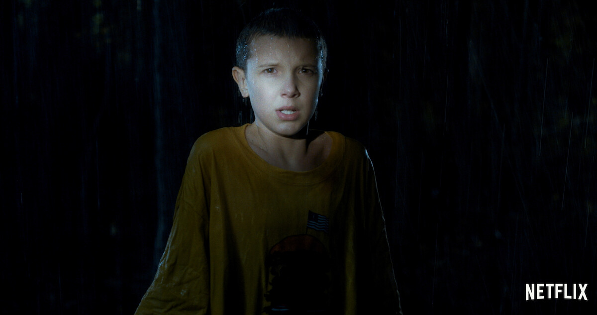 Eleven (Millie Bobby Brown) with short hair, wearing a yellow T-shirt, stands in the rain at night looking anxious.
