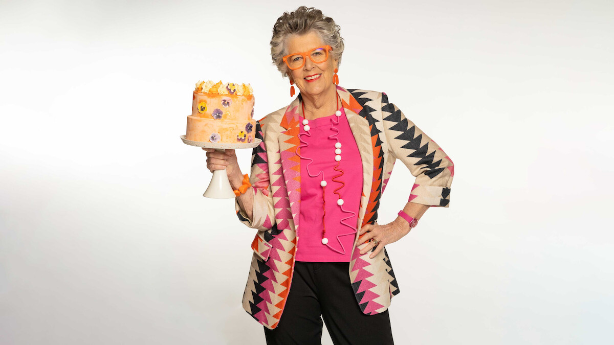 Older woman with gray hair, wearing colorful geometric jacket and glasses, stands in a bright studio holding a decorated cake on a stand, with a confident pose and cheerful expression against a plain white background.