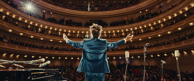 Jon Batiste stands in front of an audience on a stage. 