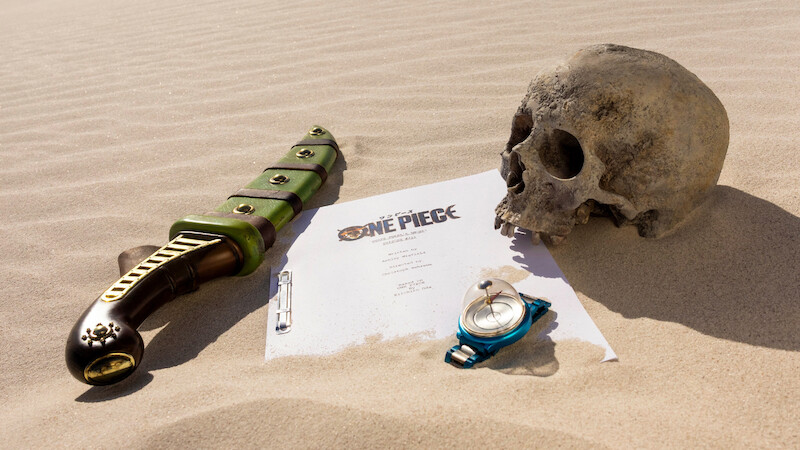 A sword, a human skull, a compass, and a ‘ONE PIECE’ script page arranged on desert sand, evoking an adventurous, mysterious, and slightly ominous atmosphere.