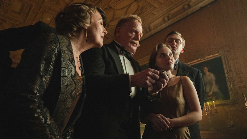 Four well-dressed people in formal evening attire stand close together in an ornate, dimly-lit room with a decorated ceiling and classical artwork, appearing to discuss something intently.