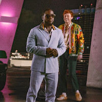 Two men standing outdoors at night, one in a light blue suit and sunglasses, the other in a colorful jacket, with city lights and modern architecture in the background, under pink and purple lighting.