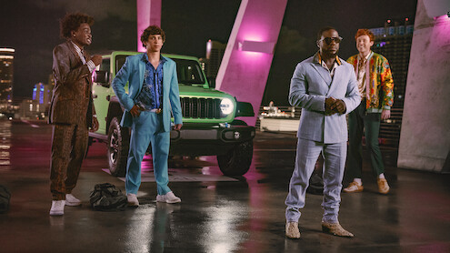 Four men in stylish suits stand in front of a green Jeep on a rooftop at night, illuminated by pink and purple lights, with a cityscape in the background.