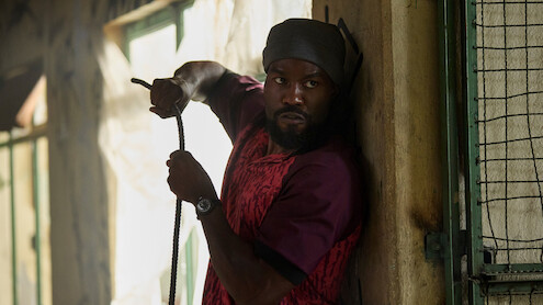 A man in a red shirt and headwrap cautiously holds a rope while pressed against a wall inside a dimly lit building with barred windows, appearing alert and wary of his surroundings.