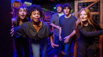 Five young adults in casual clothing look excited and curious in a colorful, dimly lit room with wooden crates and barrels, suggesting an escape room or adventure game environment.