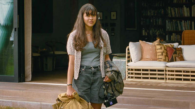 Young woman with long hair stands outside a cozy, modern house holding a coat, bag, and camera. The relaxed setting features a wicker sofa and bookshelves, evoking a calm, casual, and welcoming atmosphere.
