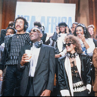 Group of musicians wearing headphones in a lively recording studio, smiling and singing together in front of a USA for Africa banner during a charity event.