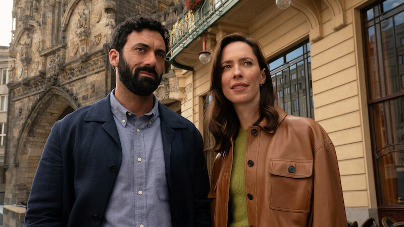 A man and woman stand outdoors in a city setting, with historic stone building details and modern architecture visible behind them. Both appear thoughtful, dressed in casual jackets, under daylight.
