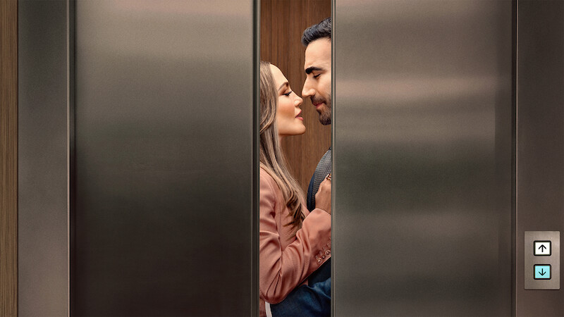 A couple standing close together inside an elevator, gazing at each other affectionately as the stainless steel elevator doors partially close, suggesting a romantic or intimate moment in a modern building setting.