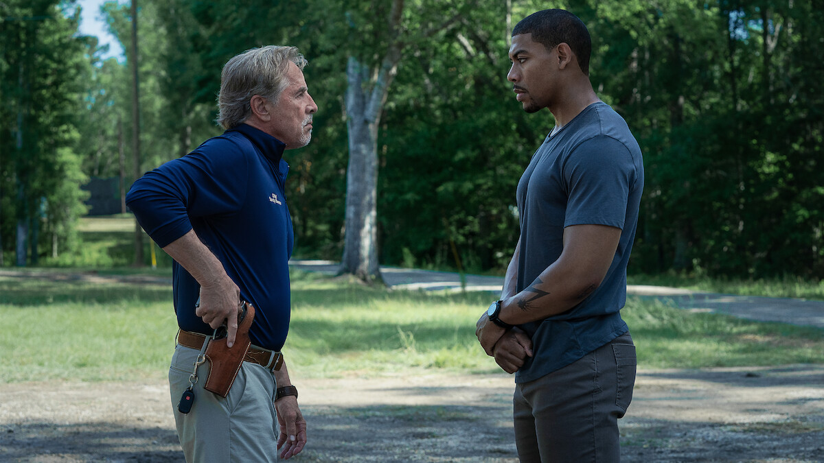 Two men stand outdoors facing each other in a grassy, wooded area. One man, older and wearing a blue shirt with khaki pants, has a hand on a holstered gun. The other, younger man, stands calmly with hands clasped.
