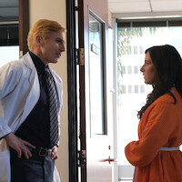 A doctor in a white coat talks to a woman in an orange sweater inside an office with large windows and a modern interior, both standing near an open door, appearing engaged in conversation.