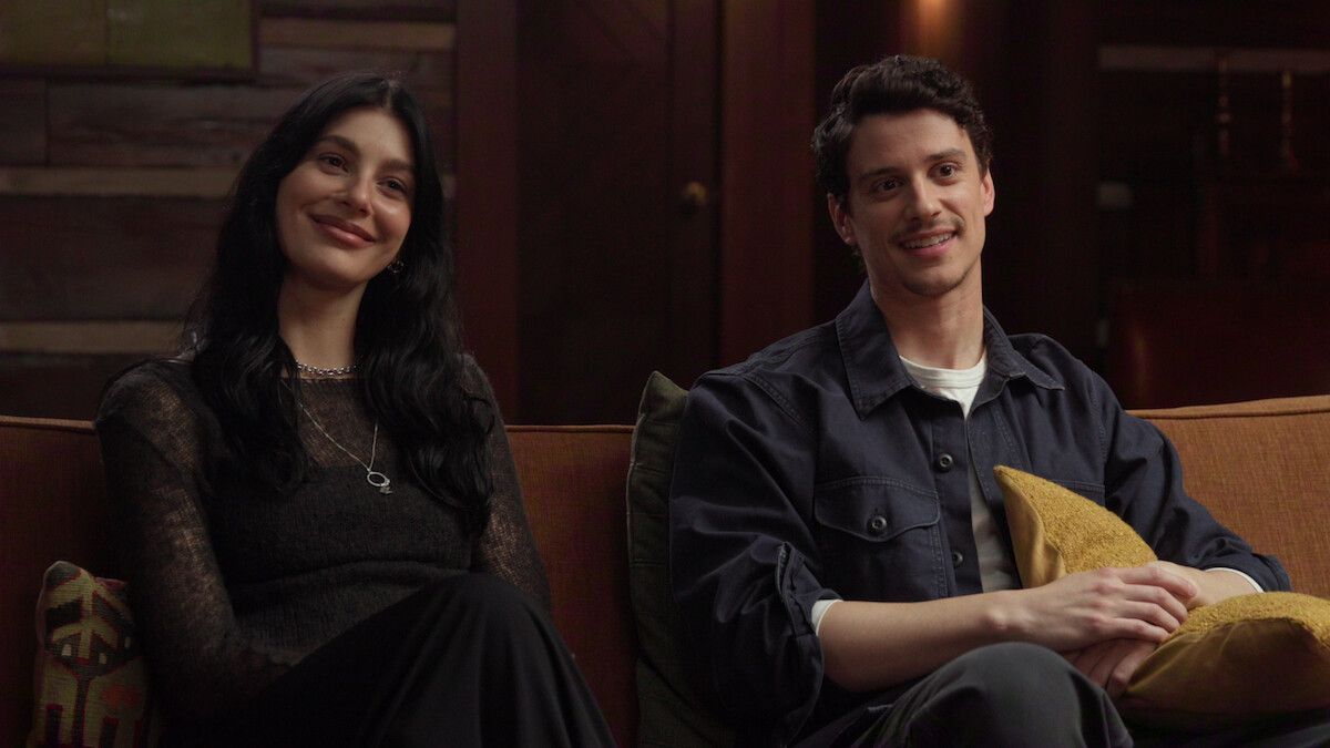 Camila Morrone and Adam DiMarco sitting on a couch in a cozy, warmly lit room with wooden walls, smiling and relaxed.