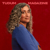 Geeta Gandbhir, director of The Perfect Neighbor, stands in front of a rich red backdrop with beautifully set curls in her graying hair, head turned toward the camera with a confident expression on her face.