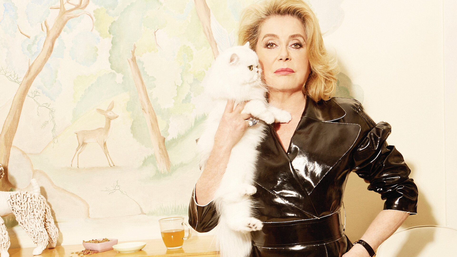 Woman in black leather outfit holding fluffy white cat, standing indoors by a painted wall with a deer mural, a table with tea, snacks, and decorative objects in a softly lit, stylish setting.