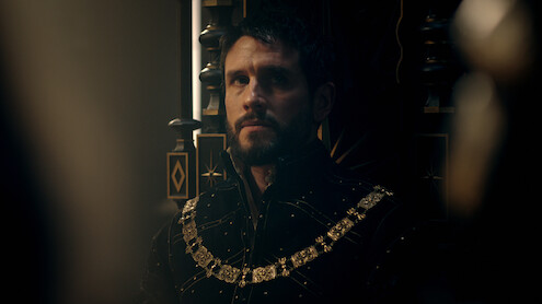 A solemn man in elaborate medieval attire with a gold chain sits on a dark, ornate throne in a dimly lit, regal setting, creating a moody, contemplative atmosphere.