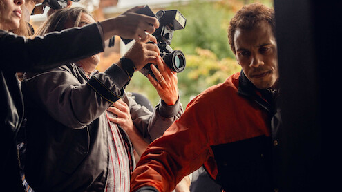 A man in a red and black jacket tries to move through a crowd outside as people with cameras and microphones surround him, suggesting a media or paparazzi situation.