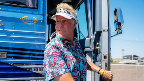 Man in a tropical shirt and visor stands at the doorway of a blue luxury bus on a sunny day, holding the door handle, with a fence and clear sky in the background.