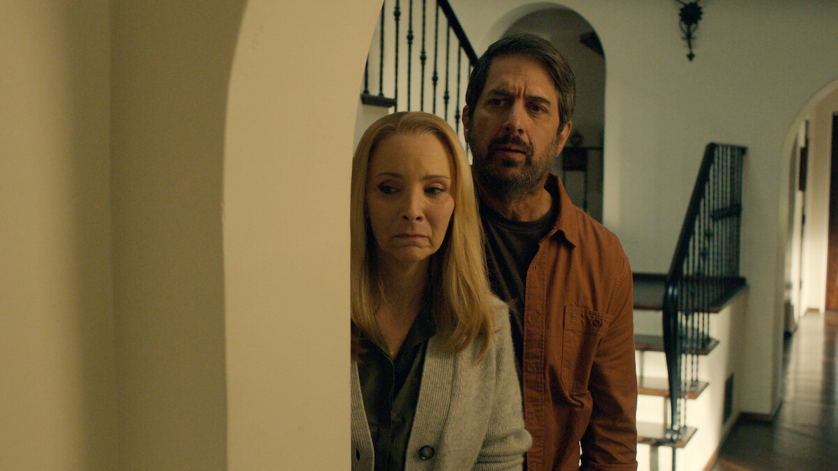 Ray Romano as Paul and Lisa Kudrow as Lydia hide behind a doorway with worried expressions in Season 1 of 'No Good Deed'