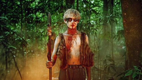 A person with painted face and fur vest stands holding a spear in a dense, misty, green forest, suggesting a primitive or survival setting.