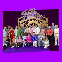 Large group of people posing together on a purple carpet in front of a golden "Wonka" gate backdrop, dressed in colorful outfits, with a purple background, suggesting a themed event or celebration setting.