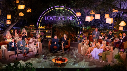 A group of well-dressed people sit in a semi-circle outdoors at night, under lanterns and trees, with a neon "Love Is Blind" sign behind them, creating a festive, lively, and glamorous TV show reunion atmosphere.