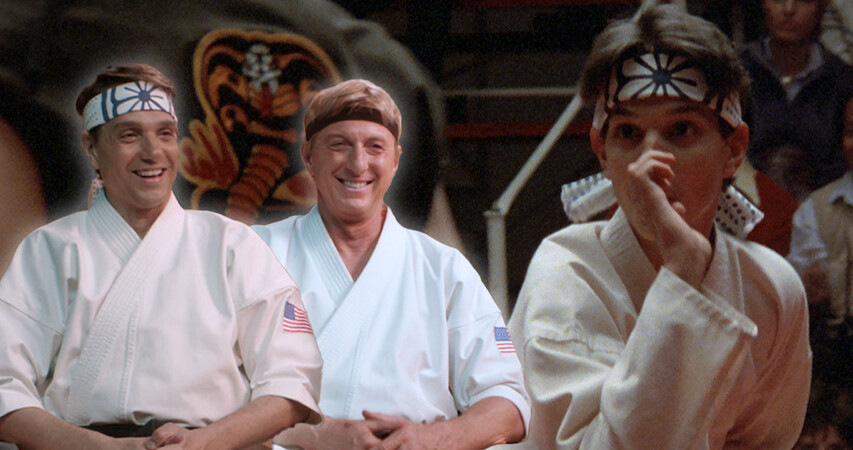 Ralph Macchio and William Zabka sit together in karate uniforms smiling looking at a young Ralph Macchio in 'The Karate Kid.'