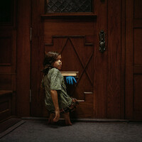 A young child kneels inside a dimly lit house by a wooden door, turning to look back as a blue-gloved hand reaches through the mail slot, creating a mysterious and suspenseful atmosphere.
