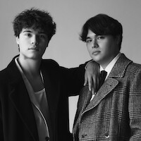 Two young men pose together in a studio, one with his arm resting on the other's shoulder. Both wear stylish coats, one in a suit and tie, in a black and white portrait with a plain background.