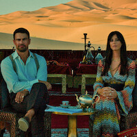 Man and woman sit at ornate table with teapot and cups in an outdoor lounge, decorated with carpets and silverware, against a desert sand dune backdrop under warm sunlight.