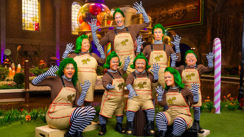 Grupo de pessoas fantasiadas de umpa-lumpas, com perucas verdes, camisas marrons, leggings listradas e macacões, no cenário de uma fábrica de doces colorida e extravagante, com guloseimas gigantes e iluminação vibrante.