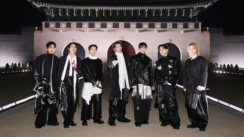 V, JIN, SUGA, RM, JUNG KOOK, J-HOPE, and JIMIN dressed in fashionable black and white outfits stand outdoors at night in front of an ornate, traditional East Asian building with arches and a large decorative roof.